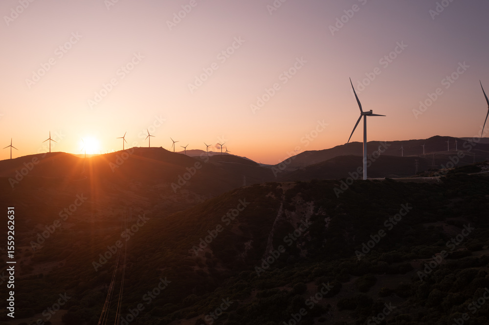 Obraz premium Wind turbines stand silhouetted against a breathtaking sunset, generating clean energy amidst rolling hills, extending to the distant horizon