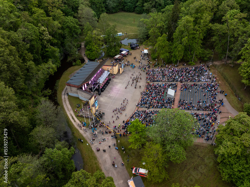 An outdoor amphitheater hosts a performance with a seated audience, surrounded by trees, pathways, a stream, and visible vehicles near the entrance.
