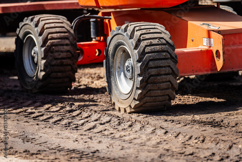 close-up of heavy construction equipment wheels on muddy site with deep tire treads and orange machine body in sunlight, industrial vehicle on dirt ground leaving visible tracks and texture patterns