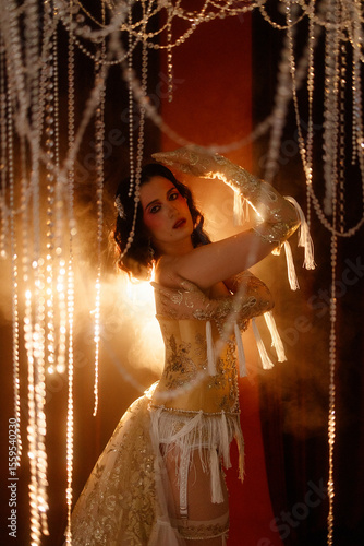 Wallpaper Mural Young Caucasian circus performer or burlesque dancer in golden vintage corset, front view, posing under chandelier on dark background. Concepts: circus, cabaret, retro, show, performance. Torontodigital.ca