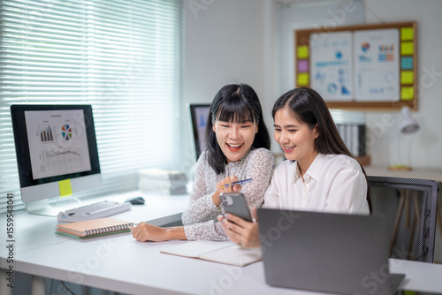 Two women are sitting at a desk with a laptop and a cell phone