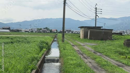  A quiet rural scene with a small canal, dirt road, and electric poles. Traditional Japanese countryside with green rice fields and distant mountains under a cloudy sky.