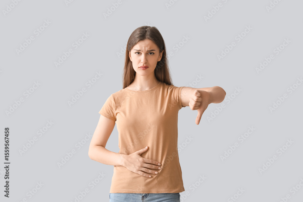 Fototapeta premium Young woman with stomach ache showing thumb-down on light background