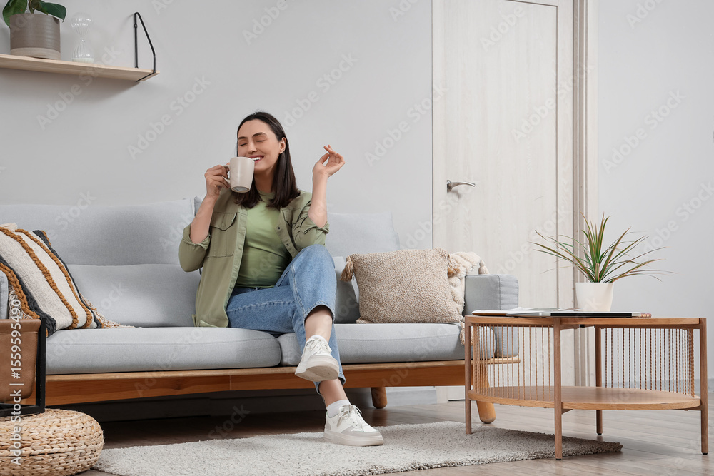 Fototapeta premium Young woman with cup of tea sitting on grey sofa near light wall at home