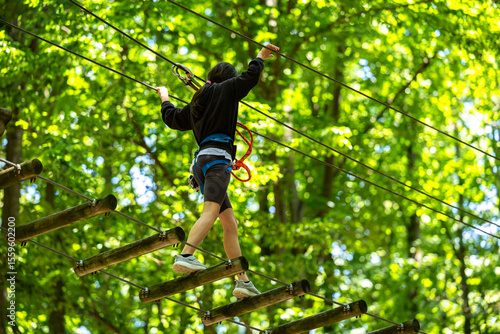 Child with harness crossing a rope bridge in a fun adventure park for kids. Outdoor forest activity focused on balance, courage, and safety. Adventure park with kids sport activities.