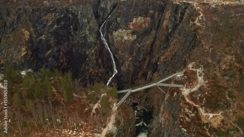 Voringsfossen waterfall cascades down rugged dark rock face with modern bridge crossing over canyon. Dramatic scale of natural wonder in Norway