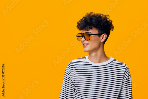 Konstfotografi Young man wearing striped shirt and stylish sunglasses looking sideways with a cheerful expression against bright orange background, showcasing modern casual fashion