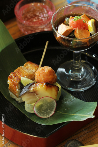 A traditional Japanese meal is beautifully arranged, featuring a variety of small bites including unagi on rice and a piece of grilled fish, served with a separate bowl of assorted delicacies on a ban
