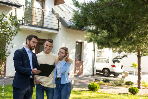 Real estate agent presenting clipboard with documents to couple in front of residential property