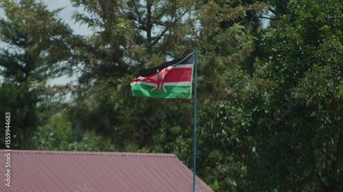 National flag flutters in wind above roof of house in settlement on Mount Elgon against backdrop of green trees