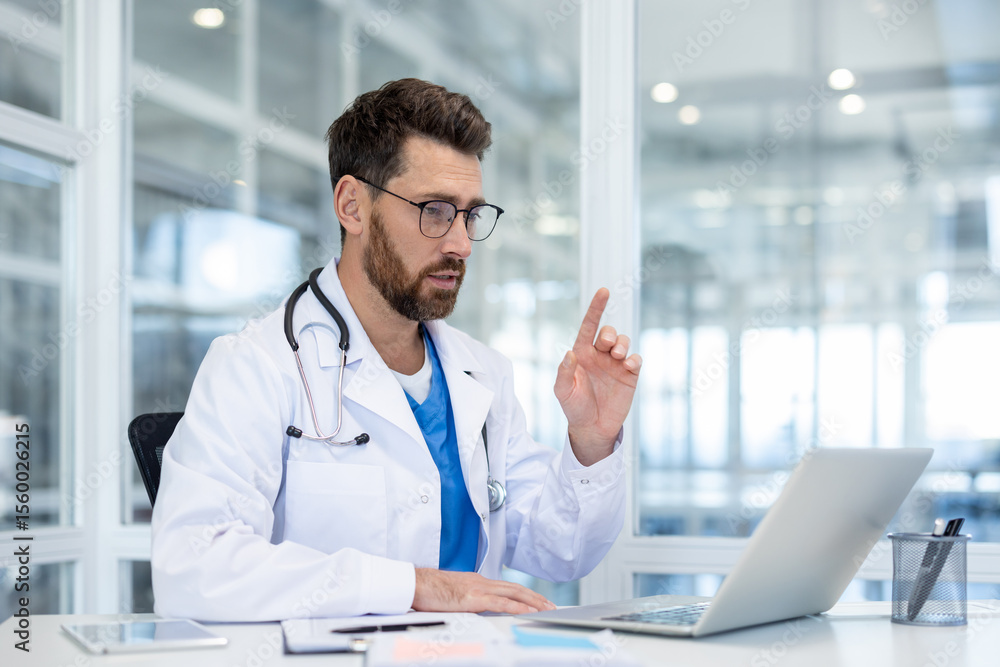 A doctor in a white coat and glasses points while looking at a laptop, likely during a telehealth consultation. A doctor in a white coat and glasses points while looking at a laptop, likely during a telehealth consultation.