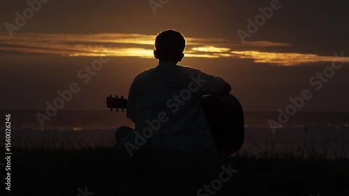 A young man sits and plays guitar on a tropical beach at sunset. A happy man enjoys the sunset while playing guitar on the beach
