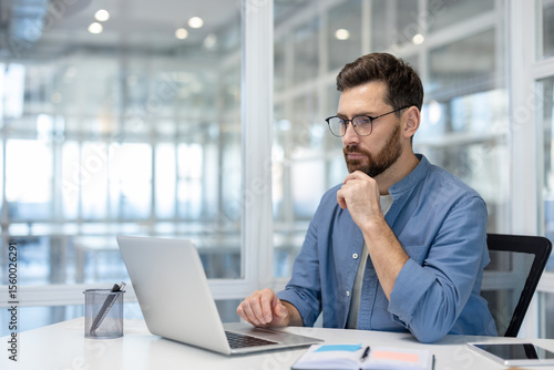 Quadro em tela A pensive man in glasses works on a laptop in a modern office setting, pondering a problem or task