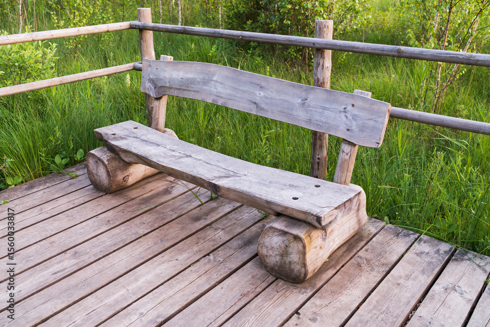 Naklejka premium Rustic handmade wooden bench on a boardwalk in a park. A tranquil spot for rest and solitude surrounded by lush greenery.