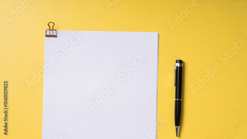 A black pen and a stack of white papers clipped with a rose gold binder clip on a solid yellow background, captured in a clean, top-down flat lay style