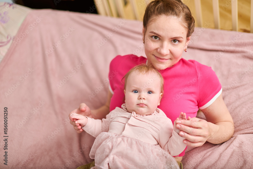 Young adult female with brown hair holding adorable baby girl in pink outfit. European ethnicity, both smiling indoors on soft pink blanket with wooden crib nearby Young adult female with brown hair holding adorable baby girl in pink outfit. European ethnicity, both smiling indoors on soft pink blanket with wooden crib nearby