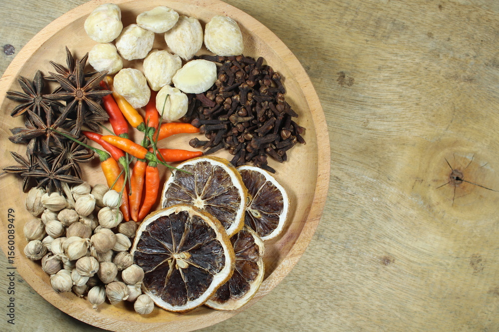 Assorted dried spices and herbs including cardamom, cloves, star anise, dried lemon, candlenut, and chili peppers arranged in a wooden bowl on rustic background. Ideal for culinary or herbal themes Assorted dried spices and herbs including cardamom, cloves, star anise, dried lemon, candlenut, and chili peppers arranged in a wooden bowl on rustic background. Ideal for culinary or herbal themes