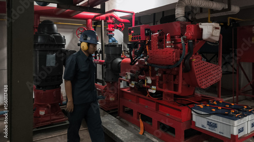 A worker in safety gear inspects a large red industrial pump and related machinery in a utility room.