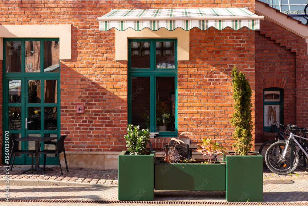 Naklejka premium Street cafe with brick walls and canopy exterior design. Front view, sunny day. An outdoor cafe with tables, chairs. Lush green plants growing in pots near house. High quality photo.