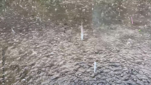 Heavy raindrops hit shallow puddles on a concrete or asphalt surface. The water's surface is filled with countless ripples, concentric circles, and tiny bubbles formed by the falling rain