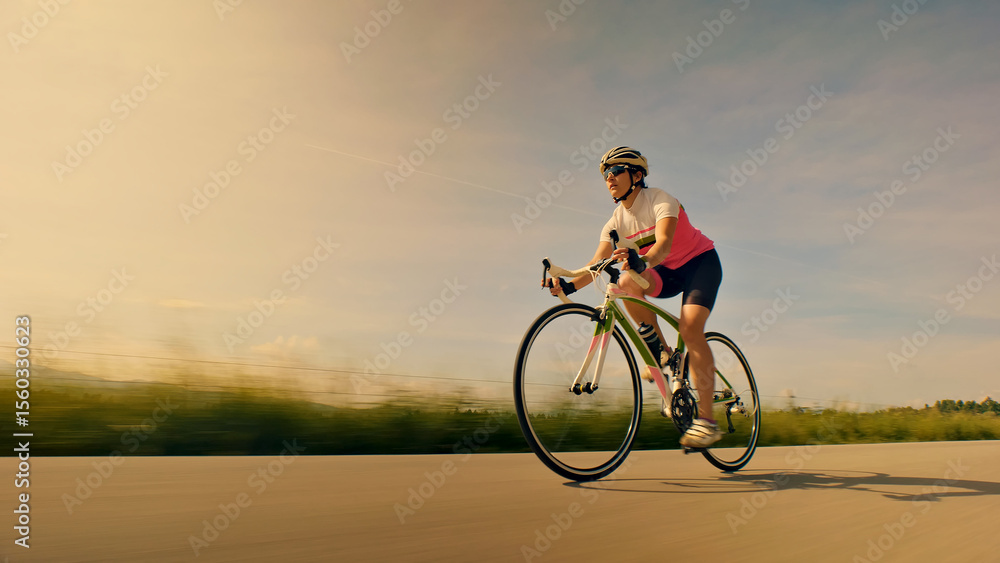 Obraz premium A woman cyclist rides her bike on an open road, wearing a helmet and sunglasses. The background features a blurred landscape, emphasizing speed and focus. Aerial view.