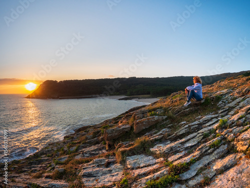 Sunset over Atlantic coast Galicia Spain woman sitting on rocky cliff facing glowing horizon peaceful bay forested hill serene moment reflection solitude freedom connection with nature