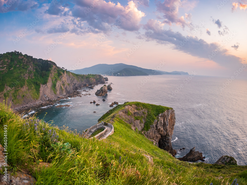 Fototapeta premium Sunset behind Gaztelugatxe Basque Country Spain winding stone bridge climbing rocky islet toward hermitage calm sea glowing sky ancient path mysticism faith solitude timeless journey