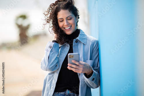 Photography Smiling woman using phone, leaning against blue wall with windblown hair