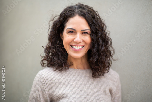 Portrait of a cheerful woman with curly hair smiling at the camera, wearing a beige sweater, standing against a plain neutral background