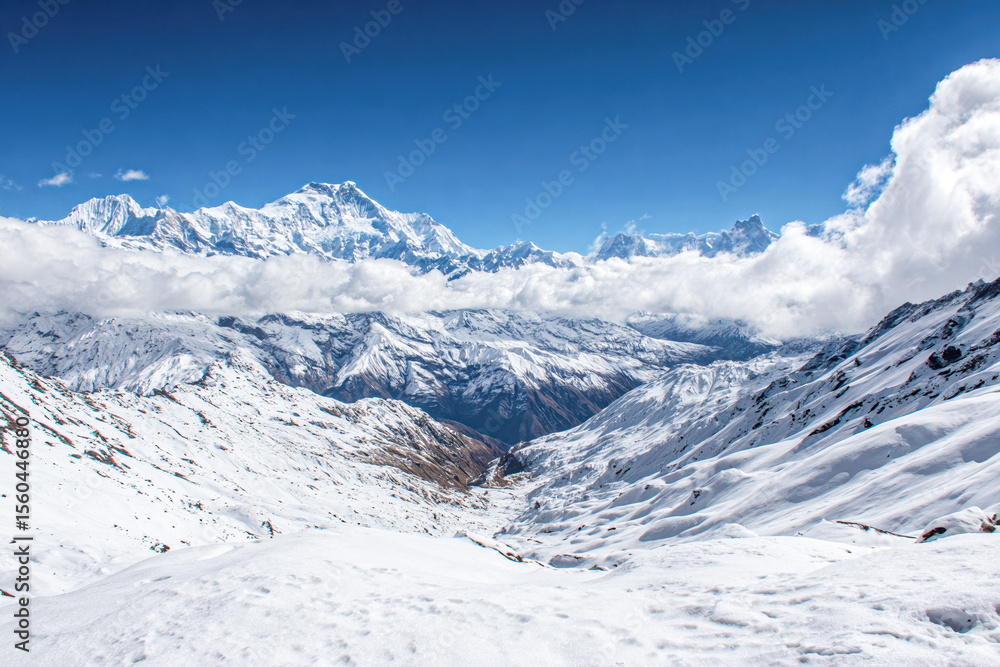 Fototapeta premium Snow-covered mountain range under a bright blue sky with scattered clouds and a valley below.