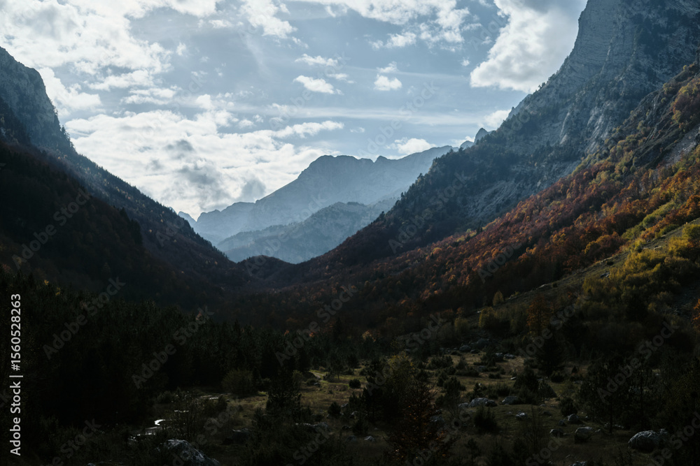 Obraz premium Dramatic view of mountain ranges in Ropojana valley during autumn at sunset in Prokletije National Park, Montenegro. Layers of colorful forest and distant peaks under cloudy sky.