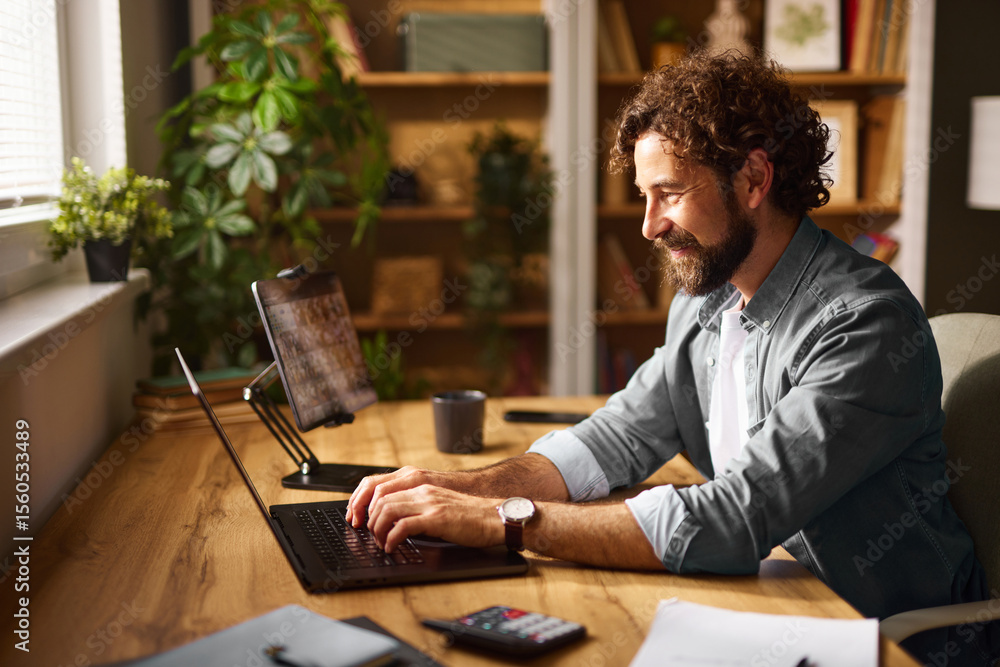 Fototapeta premium A man focuses on his laptop in a comfortable home office, surrounded by plants and wooden shelves, enjoying the warm afternoon light filtering through the window.