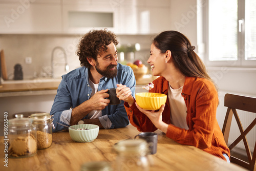 A couple sits at a wooden table in a well-lit home office, sharing a light breakfast with smiles, enjoying each other's company and a moment of connection.