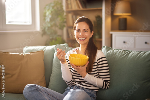 Canvas Print A woman with long dark hair smiles as she eats cereal from a yellow bowl while sitting comfortably on a couch in a well-lit, inviting living room space