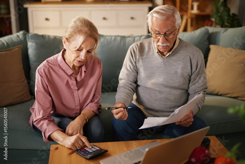 An older man and woman sit on a sofa at home, examining paperwork and using a calculator and laptop. They appear to be managing their finances or discussing insurance options.