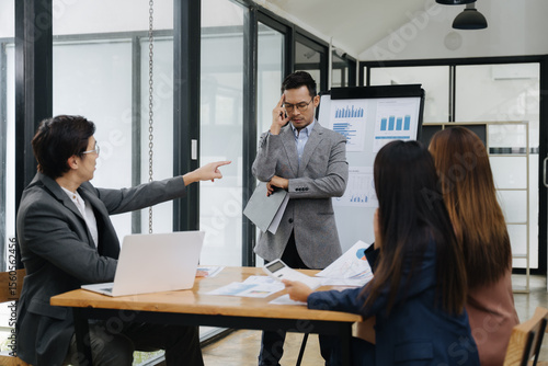 Conflict and Decisions in Boardroom: The photo captures a tense moment in a modern office, with a man pointing his finger at a colleague during a business meeting, and others engrossed in documents.
