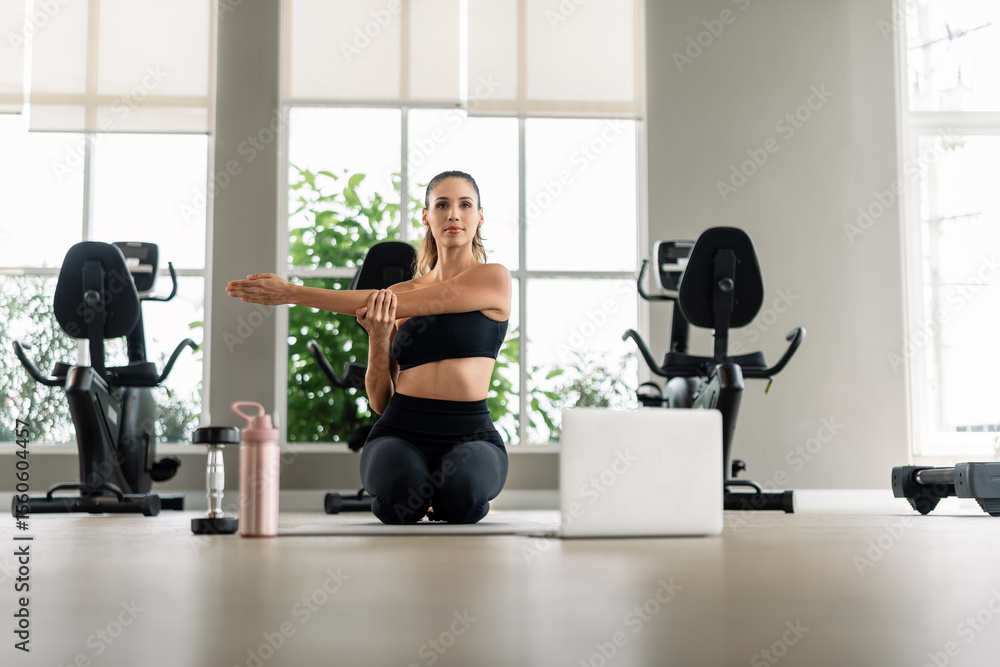 Fototapeta premium A woman is doing yoga in a gym. She is wearing a black tank top and black pants. There is a laptop on the floor in front of her