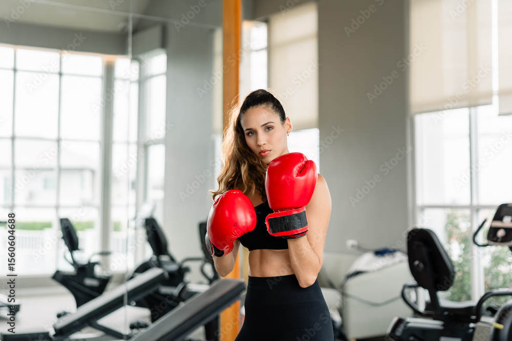 Fototapeta premium A woman wearing a red boxing glove. She is standing in front of a mirror. There are two exercise bikes in the background