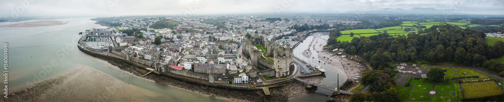 Fototapeta premium Aerial panoramic view of Caernarfon, North Wales, showcasing Caernarfon Castle, the historic town centre, and the surrounding Menai Strait and lush countryside under overcast skies