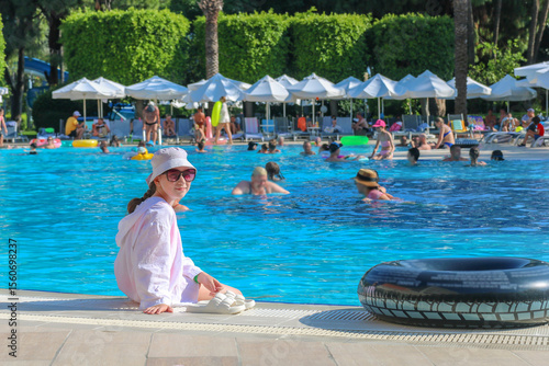 Kid girl in panama sits on edge of resort hotel pool with legs dangling in water