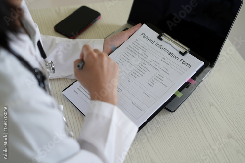 Young asian woman doctor in white uniform with stethoscope taking notes, using laptop, mobile phone, writing in medical journal, professional therapist practitioner filling documents or patient card.