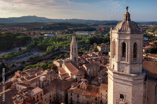 An aerial view of Girona's historic city center, featuring the cathedral, the Onyar River winding through colorful buildings, and ancient city walls amidst lush hills