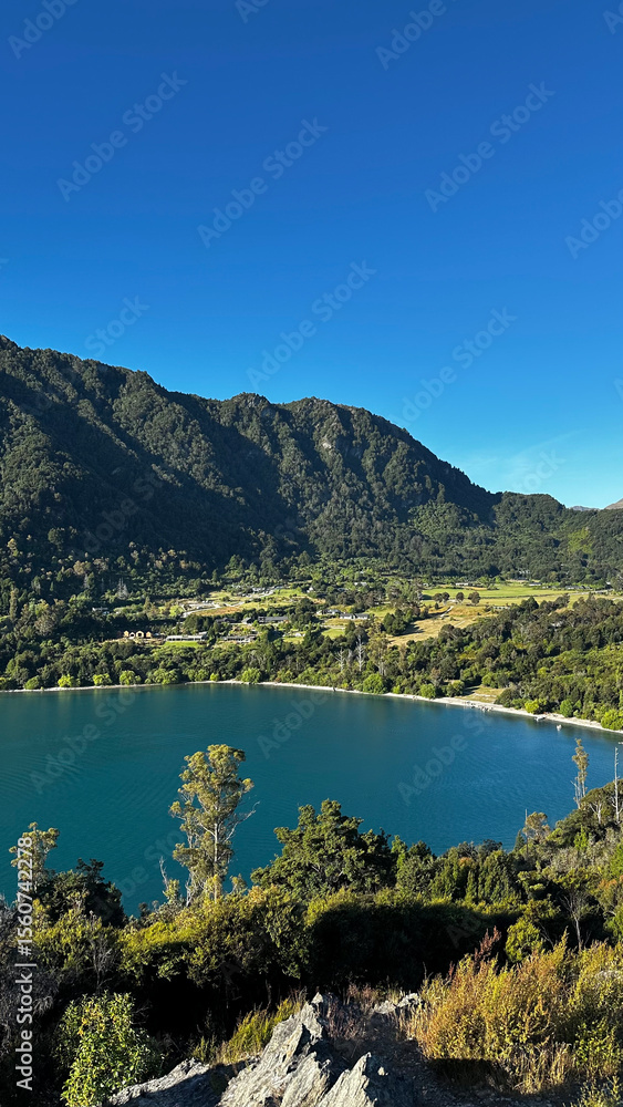 Obraz premium Scenic view of a clear blue lake surrounded by green forest and mountains under a bright blue sky in Queenstown, New Zealand