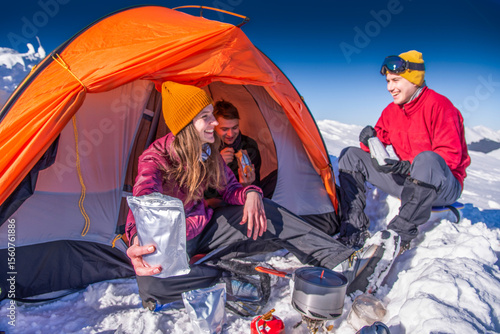 Smiling hikers enjoying winter camping in snowy mountains. Woman holding freeze-dried food pouch outside tent