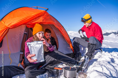 Female hiker showing freeze-dried food pouch near tent in snowy mountains. Group of campers eating and resting during winter expedition