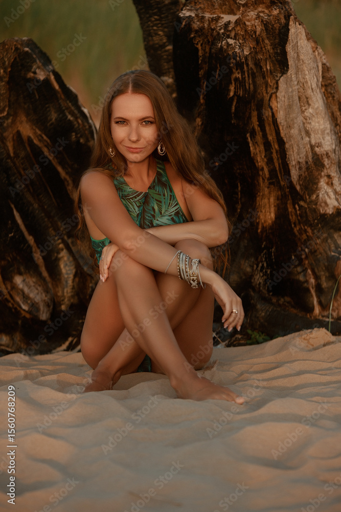 Naklejka premium A woman in a green palm-print bikini, adorned with jewelry, gazes softly into the distance, illuminated by the warm glow of the setting sun