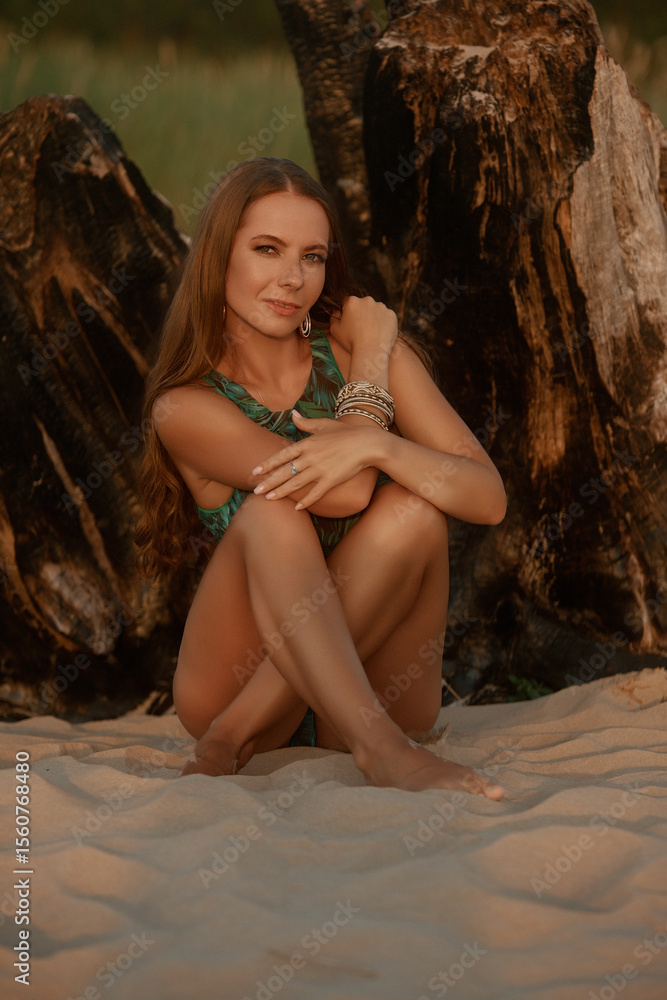 Naklejka premium A woman in a green palm-print bikini, adorned with jewelry, gazes softly into the distance, illuminated by the warm glow of the setting sun