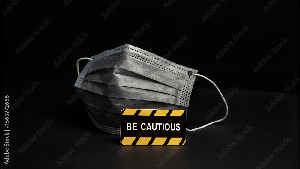 Naklejka premium A protective face mask is placed beside a sign that reads be cautious against a dark background, urging vigilance