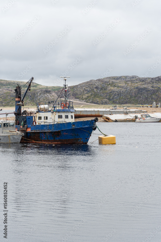 Fototapeta premium Old blue fishing vessel anchored near the shore with rugged hills in the background and cloudy sky above