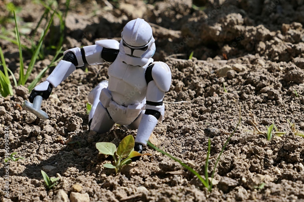 Naklejka premium Star Wars figure of Clone Trooper digging out some weed plant from garden soil, while kneeling on ground. Spring daylight sunshine. 